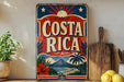 A vintage-style sign with the text "COSTA RICA" displayed prominently, featuring a tropical landscape with palm trees, mountains, and a sunburst design. The sign is placed on a shelf next to a wooden cutting board and a potted plant, creating a rustic and inviting atmosphere.