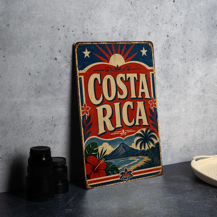 A vintage-style metal sign with the text "COSTA RICA" prominently displayed, featuring a tropical landscape with palm trees, mountains, and a sun. The sign is placed on a gray surface, with a stack of black camera lenses and a white dish visible in the background.