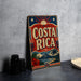 A vintage-style metal sign with the text "COSTA RICA" prominently displayed, featuring a tropical landscape with palm trees, mountains, and a sun. The sign is placed on a gray surface, with a stack of black camera lenses and a white dish visible in the background.