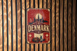 A vintage metal sign with the word "DENMARK" prominently displayed, featuring a red background with a sunburst design and various landmarks.