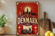 A vintage-style metal sign with a red and white color scheme, featuring a depiction of Denmark and its landmarks, such as windmills and a castle, along with decorative elements like stars and flowers.