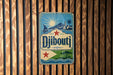 A vintage-style metal sign with the word "Djibouti" displayed prominently, featuring a sailboat, palm trees, and a mountain landscape.