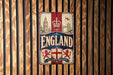 A vintage metal sign with the word "ENGLAND" prominently displayed, featuring a red and white color scheme and a shield with a lion and a crown.
