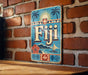 A vintage-style metal sign with the word "Fiji" prominently displayed, featuring a tropical island landscape with palm trees, a sailboat, and a hibiscus flower.