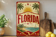 A vintage-style sign with the word "FLORIDA" prominently displayed, featuring palm trees, a sailboat, and a beach scene. The sign is placed on a shelf next to a wooden cutting board and some kitchen utensils, with a potted plant and a lemon visible in the background.