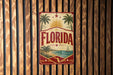 A vintage-style metal sign with the word "FLORIDA" prominently displayed, featuring palm trees, a sun, and a sailboat, all set against a tropical beach backdrop.