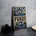 a vintage-style metal sign with a blue and white color scheme, displaying the word "FUKUI" prominently. The sign also includes a small illustration of a castle and a wave, adding to its decorative design. The sign is placed on a gray countertop, with a stack of black camera lenses and a white plate visible in the background.