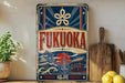 a vintage-style sign for Fukuoka, with a blue and red color scheme and a Japanese-style illustration of a temple and waves. The sign is displayed on a shelf next to a wooden cutting board and some potted plants.
