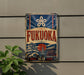 a vintage-style sign with the word "FUKUOKA" prominently displayed, along with a Japanese temple and other cultural elements. The sign is mounted on a wall, and there is a plant visible to the left of the sign.