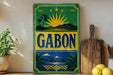 a vintage-style sign with a tropical design, including palm trees, mountains, and a sun, along with the word "GABON" prominently displayed.