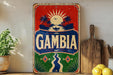 a vintage-style sign with the word "Gambia" prominently displayed, surrounded by tropical elements such as palm trees and a sun. The sign is placed on a wooden surface, with a potted plant and a wooden cutting board visible in the background.