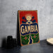 A vintage-style sign with the word "GAMBIA" prominently displayed, featuring a tropical landscape with palm trees and a sun.