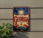 A vintage-style metal sign with the word "Georgia" prominently displayed, featuring a sun and stars design, against a gray wall.