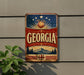 A vintage-style metal sign with the word "GEORGIA" prominently displayed, featuring a peach, sun, and mountain landscape.