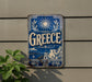 a blue and white sign with the word "Greece" prominently displayed, along with a sun and olive tree design.