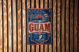 A vintage-style metal sign with the word "GUAM" prominently displayed, featuring a palm tree, sun, and other tropical elements.