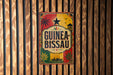 A vintage-style metal sign with the text "GUINEA-BISSAU" displayed on a wooden wall.
