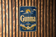 a blue and gold sign with the word "Gunma" on it, mounted on a wooden wall.