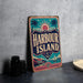 A vintage-style metal sign with the text "HARBOUR ISLAND" is displayed on a table, accompanied by a stack of black camera lenses and a white dish.