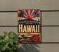 A vintage-style metal sign with the word "HAWAII" prominently displayed, featuring a tropical landscape with palm trees, a sun, and a wave.