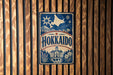 A vintage-style metal sign with the word "Hokkaido" prominently displayed, featuring a star and snowflake design, and a depiction of a building and landscape.