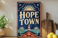 A vintage-style sign for "Hope Town" with a lighthouse and palm trees, placed on a shelf next to a wooden cutting board and some lemons.