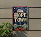 A vintage-style sign for "Hope Town" mounted on a wall, featuring a lighthouse, palm trees, and a sun.