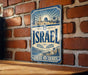 a vintage-style metal sign with the word "ISRAEL" prominently displayed, hanging on a brick wall.