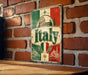 A vintage-style metal sign with the word "Italia" prominently displayed, featuring a combination of green, red, and white colors, along with images of landmarks and scenery associated with Italy.