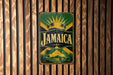 A vintage-style metal sign with the word "Jamaica" prominently displayed, featuring a tropical landscape with palm trees and a sunburst design.