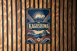 A vintage-style metal sign with the word "KAGOSHIMA" prominently displayed, featuring a sun and mountain landscape design.