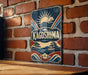 A vintage-style metal sign with the word "KAGOSHIMA" prominently displayed, featuring a sun and mountain landscape design.