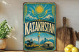a vintage-style metal sign with the word "Kazakhstan" prominently displayed, along with a sun and mountains in the background. The sign is placed on a wooden surface, with a potted plant, a wooden cutting board, and some lemons nearby.