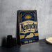 A vintage-style metal sign with the word "Kentucky" prominently displayed, featuring a sun and bridge design, is placed on a gray countertop next to some black camera lenses.