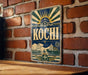 A vintage-style metal sign with the word "Kochi" prominently displayed, featuring a sunburst design and various decorative elements.