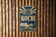 A vintage-style metal sign with the word "Kochi" prominently displayed, featuring a sunburst design and various nautical elements.