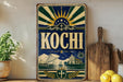 A vintage-style metal sign with the word "Kochi" prominently displayed, featuring a sunburst design and various nautical elements such as a ship, palm tree, and building. The sign is placed on a shelf next to a wooden cutting board and a potted plant, creating a nautical-themed display.