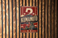 A vintage-style metal sign with the word "KUMAMOTO" prominently displayed, featuring a Japanese temple and mountain in the background.