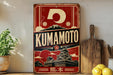 A vintage-style metal sign with the word "KUMAMOTO" prominently displayed, featuring a Japanese temple and mountain landscape. The sign is placed on a shelf next to a wooden cutting board and some potted plants.