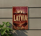 A vintage-style metal sign with the word "LATVIA" prominently displayed, featuring a scenic landscape with a sunburst effect and silhouetted buildings.