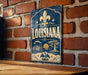 A vintage-style metal sign with the word "LOUISIANA" prominently displayed, featuring a fleur-de-lis symbol and decorative elements.