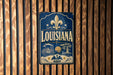 A vintage-style metal sign with the word "LOUISIANA" prominently displayed, featuring a fleur-de-lis symbol and various decorative elements.