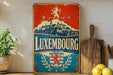 A vintage-style metal sign with the text "LUXEMBOURG" prominently displayed, featuring a castle and a lion emblem. The sign is placed on a shelf next to a wooden cutting board and a potted plant.
