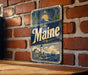 A vintage Maine-themed metal sign is displayed on a brick wall, featuring a lighthouse, mountains, and a sun.