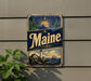 A vintage Maine beer sign is displayed on a wall, featuring a lighthouse, mountains, and a sunset.