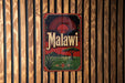 A vintage metal sign with the word "Malawi" displayed prominently in the center, featuring a sun-like design and a landscape with a tree and mountains.