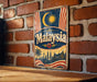 A vintage metal sign with the word "Malaysia" prominently displayed, featuring a crescent moon and sun symbol, a red and white striped pattern, and a depiction of a city skyline.