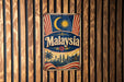 A vintage metal sign with the word "Malaysia" prominently displayed, featuring a crescent moon, sun, and a flag-like design.