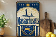 A vintage-style metal sign with the text "Massachusetts" prominently displayed, featuring a lighthouse and sailboat illustration. The sign is placed on a wooden surface, with a potted plant, a wooden cutting board, and some lemons nearby.
