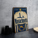 A vintage metal sign with the text "Massachusetts" and a lighthouse illustration, placed on a gray countertop next to some black camera lenses.